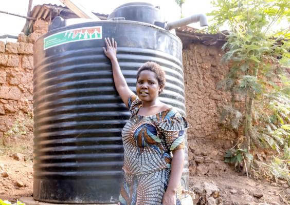 Nyirahabimana beside the household water tank installed through improved investments and planning, reflecting the family's growing resilience and improved living conditions. @FAO Rwanda/Christian Ngabonziza