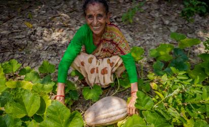 An elderly woman smiling while squatting in a garden. She is surrounded by green plants, and in front of her is a large, pale pumpkin-like vegetable.