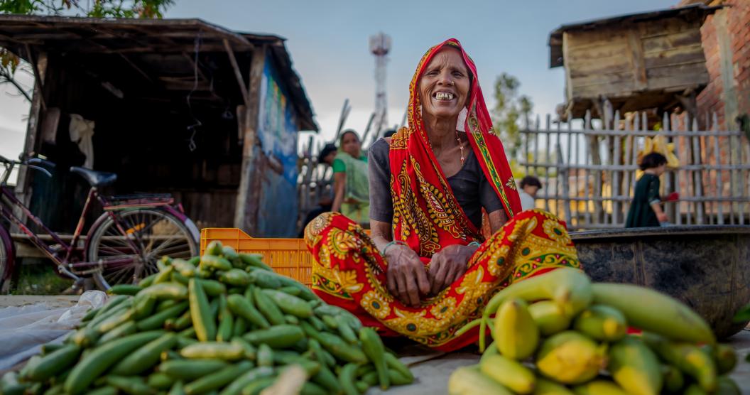 A woman in a red and orange saree sits infront of two piles of vegetables.